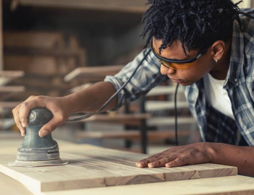 A young male carpenter sanding down a plank of wood with an electric sander in his workshop.