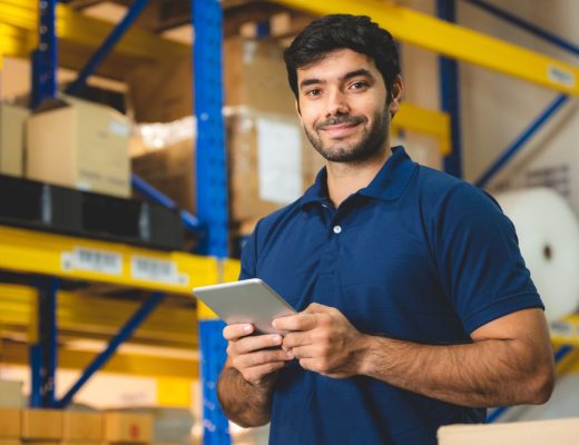 A young man smiling at the cameral while standing in a warehouse and holding a tablet.