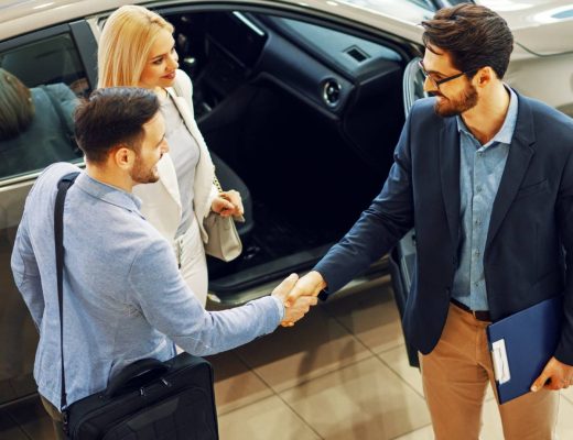 A car consignment agent and a young couple shake hands beside the open door of a shining car in a showroom.