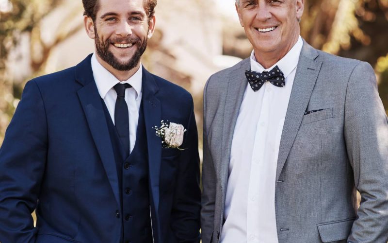 Two man are standing next to each other. The young man is wearing a dark blue suit. The old man has a gray suit and bowtie.