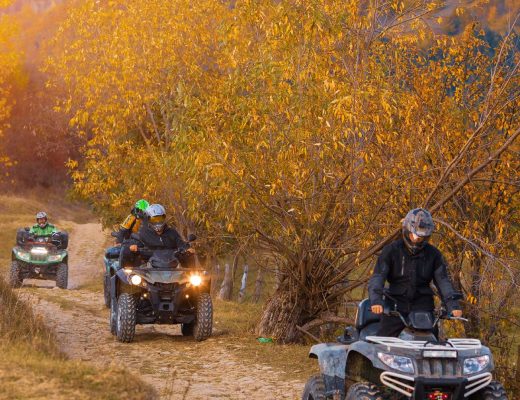 Four people wearing helmets ride their individual ATVs on a dirt trail surrounded by trees with yellow leaves.