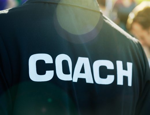 A close-up of a sports coach wearing a black T-shirt with the word “coach” written on the back in white letters.