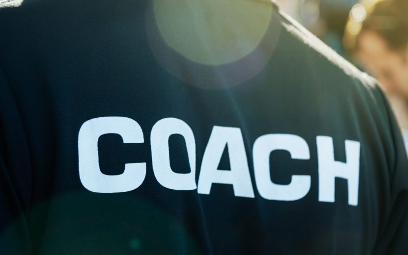 A close-up of a sports coach wearing a black T-shirt with the word “coach” written on the back in white letters.