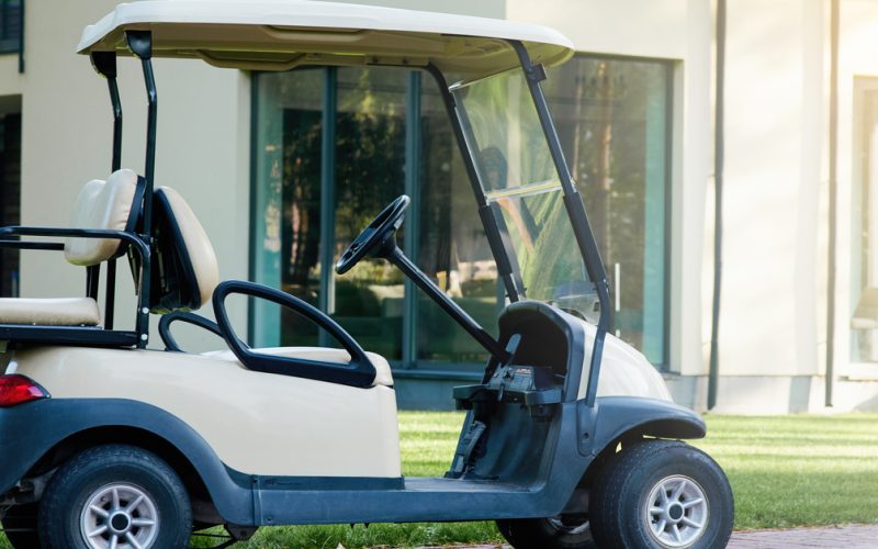 A cream and black golf cart parked outside of a nice house in the suburbs. It has extra seating in the back.