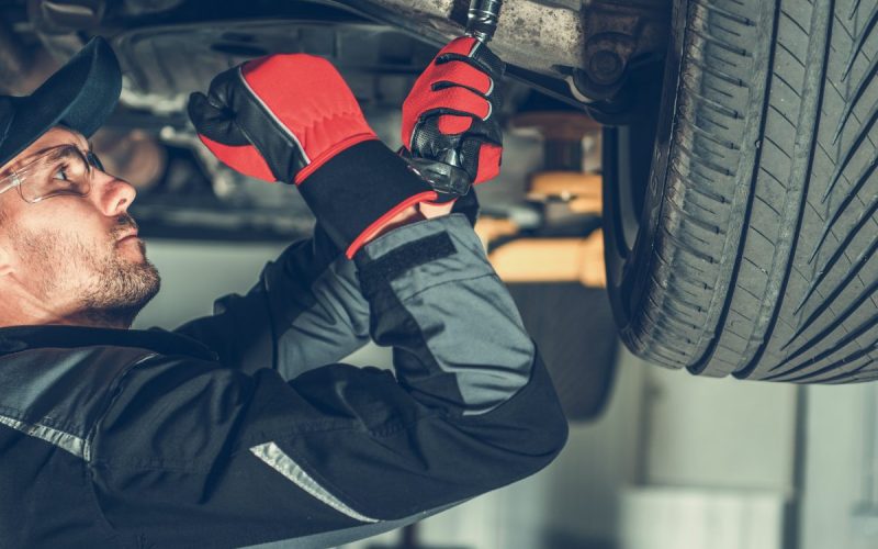 A mechanic stands beneath a vehicle, working on the suspension. He is wearing gloves and protective glasses.