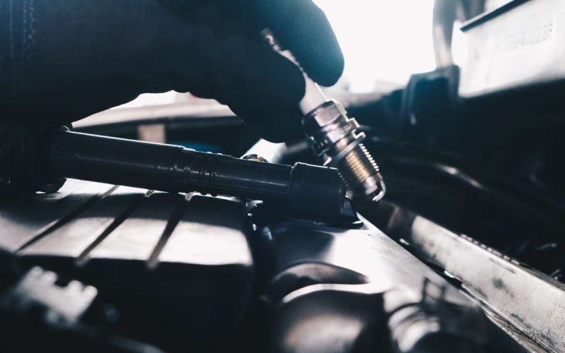 A close-up of a mechanic's gloved hand holding a brand-new spark plug over a vehicle's engine compartment.