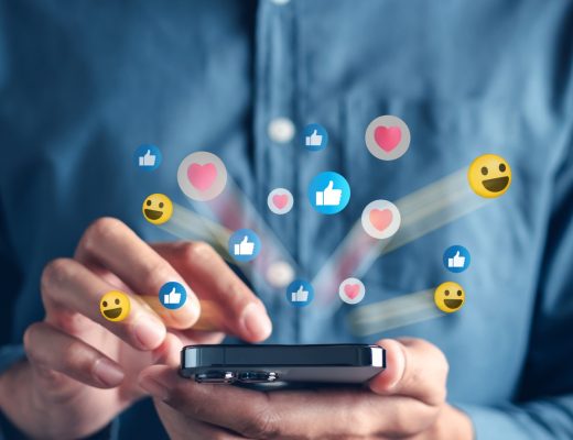 A man in a chambray dress shirt holding a black phone and tapping it as emoji icons fly outward from the screen.