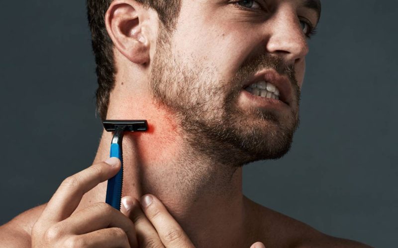 A man grimacing as he pulls the skin of his neck taut to shave a patch of beard on red, irritated skin.