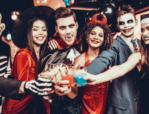 A group of six friends dressed in spooky Halloween costumes, grinning as they cheers with party decorations around.