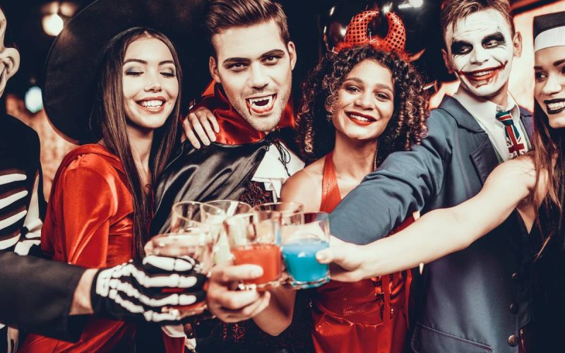 A group of six friends dressed in spooky Halloween costumes, grinning as they cheers with party decorations around.