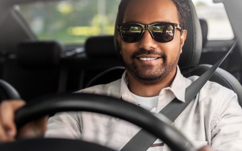 A man wearing black sunglasses and a white shirt is driving a car. He has his hands on the steering wheel.