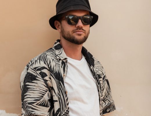 A young man in a white t-shirt and open, short-sleeved button-down with palm leaves wears sunglasses and a bucket hat.