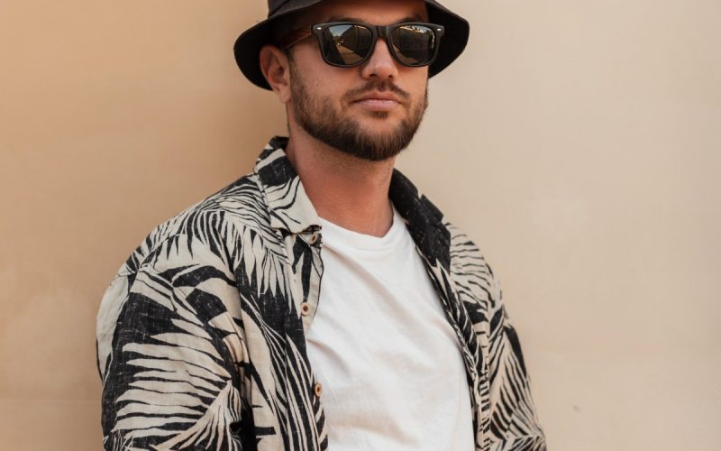 A young man in a white t-shirt and open, short-sleeved button-down with palm leaves wears sunglasses and a bucket hat.