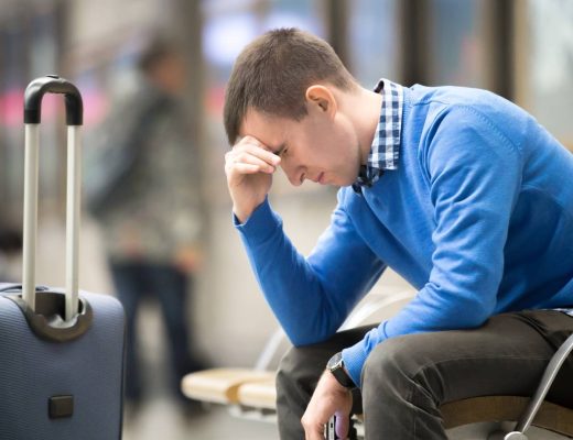 A young, frustrated man sitting in the airport. He’s holding his head while looking at the floor. His suitcase is near him.