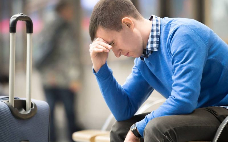 A young, frustrated man sitting in the airport. He’s holding his head while looking at the floor. His suitcase is near him.