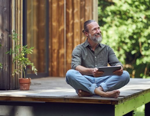 A bearded man holding a tablet while sitting on the deck of his tiny home. He’s smiling and has his legs crossed.