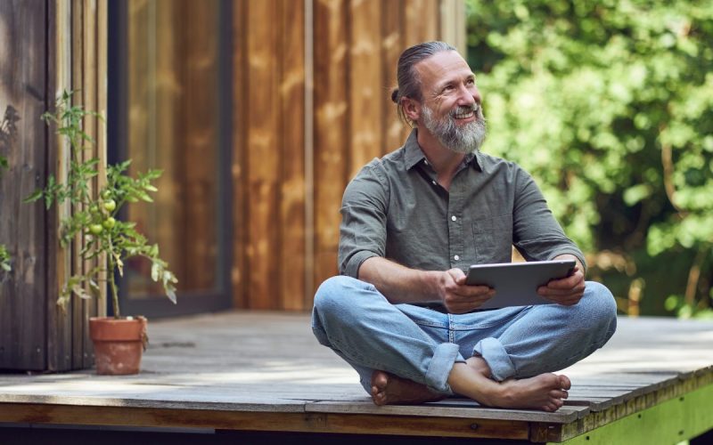 A bearded man holding a tablet while sitting on the deck of his tiny home. He’s smiling and has his legs crossed.