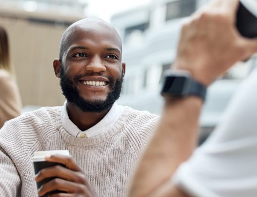 A male employee in a neutral sweater sitting in an outdoor employee break area and enjoying coffee with others.