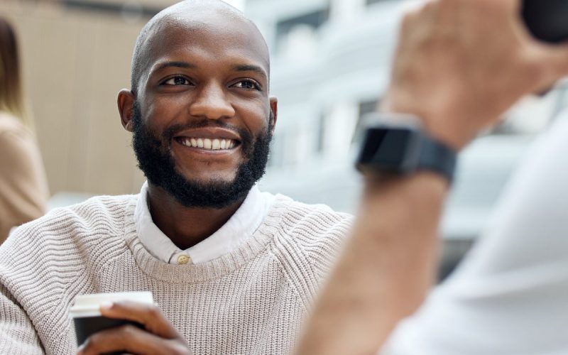 A male employee in a neutral sweater sitting in an outdoor employee break area and enjoying coffee with others.