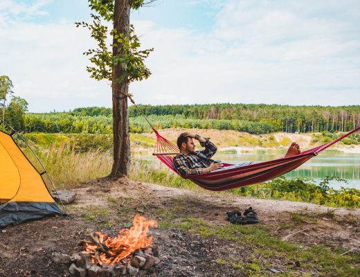 A man in a hammock strung between two trees looks out at a lake. A yellow tent and campfire are set up behind him.