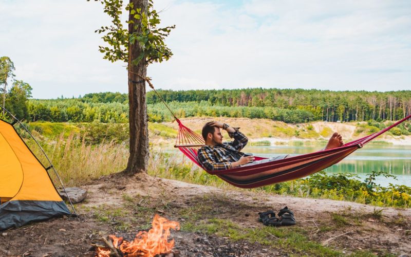 A man in a hammock strung between two trees looks out at a lake. A yellow tent and campfire are set up behind him.