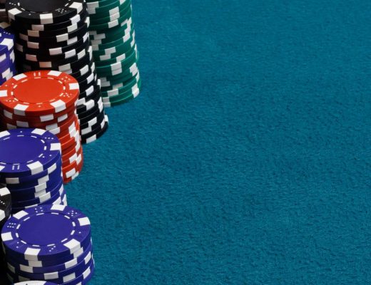 Stacks of gambling chips on a teal-colored poker table felt. The chips are blue, red, white, green, and black.