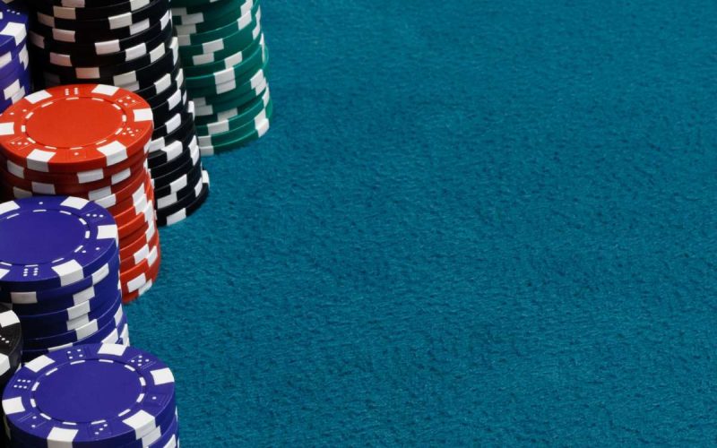 Stacks of gambling chips on a teal-colored poker table felt. The chips are blue, red, white, green, and black.