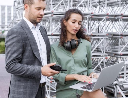 Two event managers stand outside near a concert stage and look at a laptop. The man holds a walkie talkie.