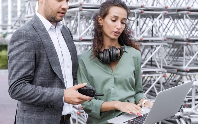 Two event managers stand outside near a concert stage and look at a laptop. The man holds a walkie talkie.