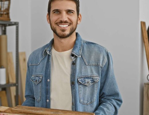 A man wearing a denim jacket and white shirt holds pieces of wood. Wood and woodworking equipment is behind him.