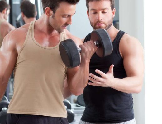 A male fitness trainer in a gym, stands by a young man, ensuring he has proper form as he does bicep curls with dumbbells.