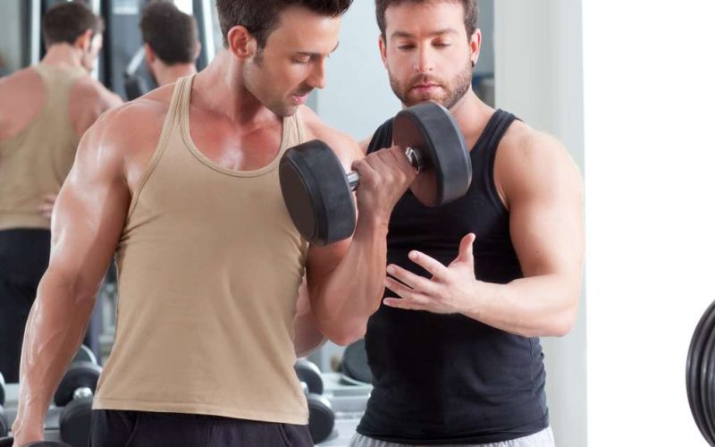 A male fitness trainer in a gym, stands by a young man, ensuring he has proper form as he does bicep curls with dumbbells.