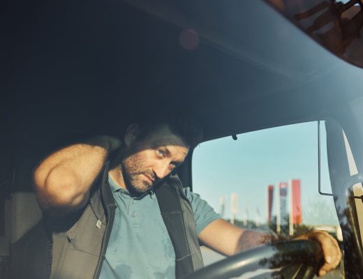 A male truck driver wearing a blue shirt and vest holds his hand to his neck in response to aching pain.