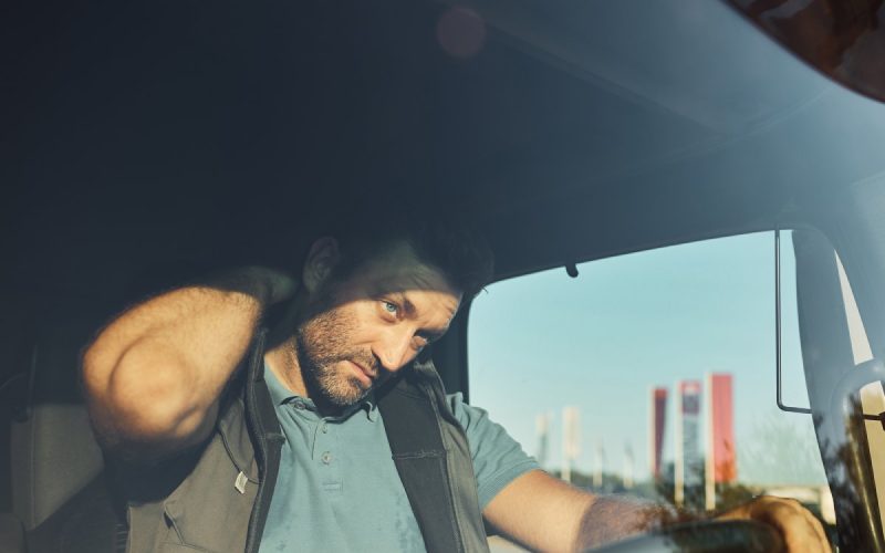 A male truck driver wearing a blue shirt and vest holds his hand to his neck in response to aching pain.