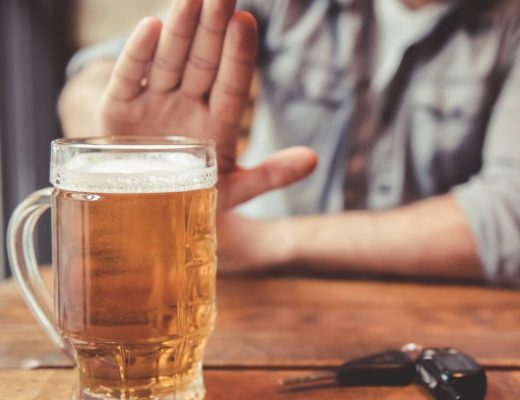 A blurred man sitting at a pub table holds out his hand to refuse an in-focus pint of beer next to a set of car keys.