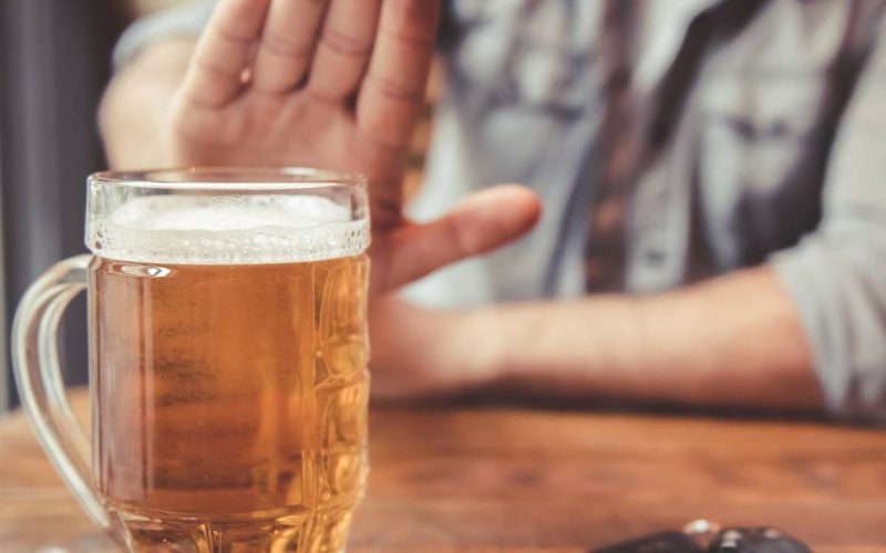 A blurred man sitting at a pub table holds out his hand to refuse an in-focus pint of beer next to a set of car keys.