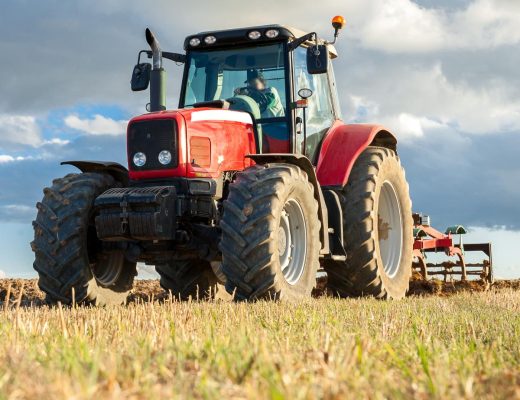 A large red tractor with a tiller driving through a field on a cloudy day. The grass is a blend of brown and green.