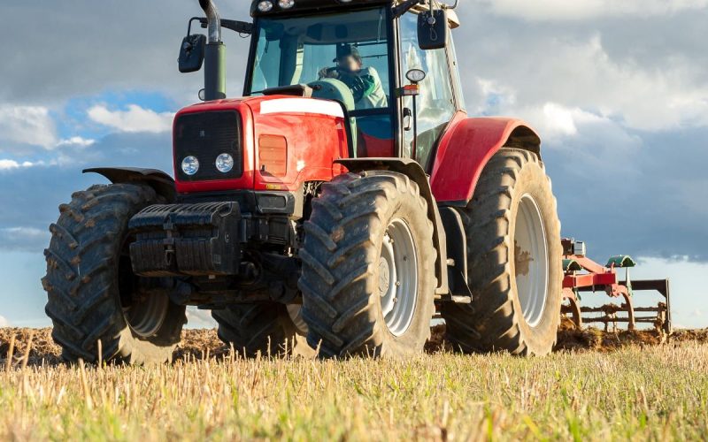 A large red tractor with a tiller driving through a field on a cloudy day. The grass is a blend of brown and green.