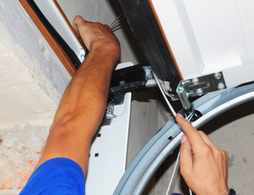 A man in a blue shirt and baseball cap is reaching up toward an open garage door. He is working on it with a screwdriver.