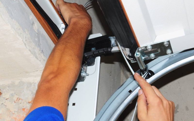 A man in a blue shirt and baseball cap is reaching up toward an open garage door. He is working on it with a screwdriver.