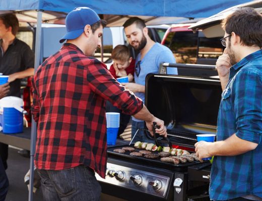 A group of men are holding blue plastic cups and enjoying an outdoor party. One man is cooking meat on a grill.