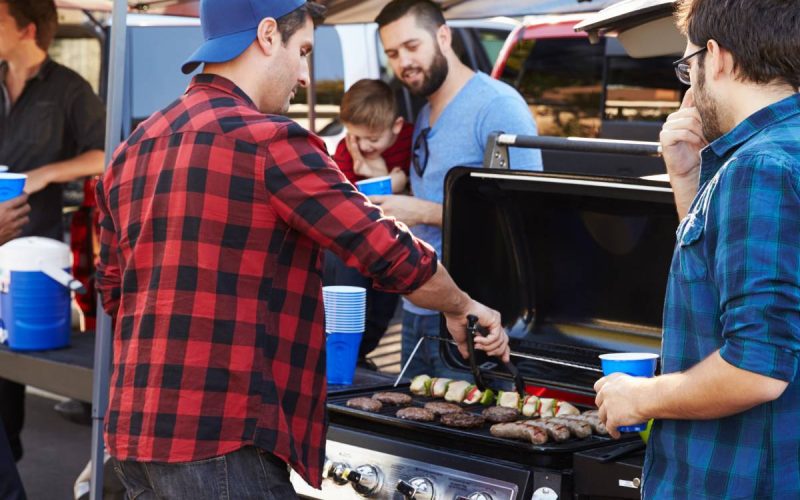 A group of men are holding blue plastic cups and enjoying an outdoor party. One man is cooking meat on a grill.