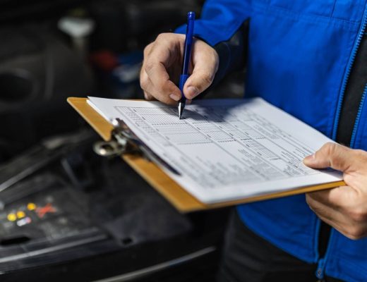 A man wearing a blue jacket, taking down notes on his paper and clipboard, stands in front of an open vehicle hood.