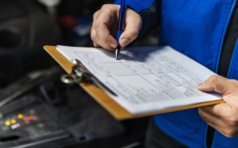 A man wearing a blue jacket, taking down notes on his paper and clipboard, stands in front of an open vehicle hood.