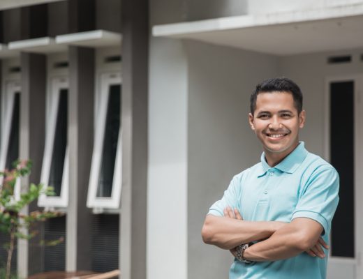 A man stands in front of a home with his arms crossed. He's wearing a blue shirt and smiling toward the camera.