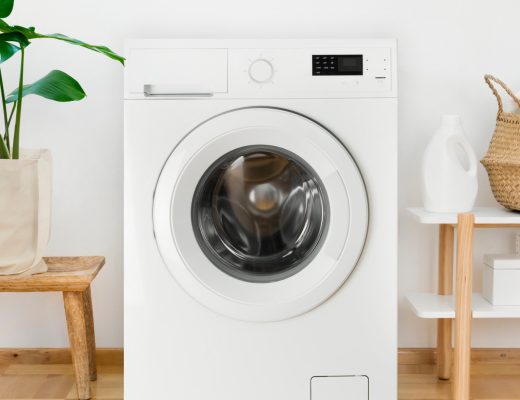 A white washing machine is sitting between a green plant and a shelf with laundry supplies and a basket.