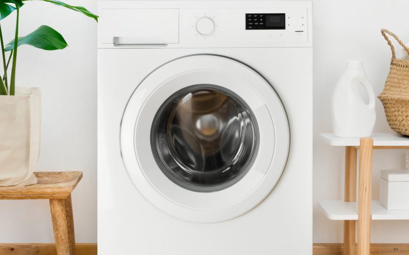 A white washing machine is sitting between a green plant and a shelf with laundry supplies and a basket.