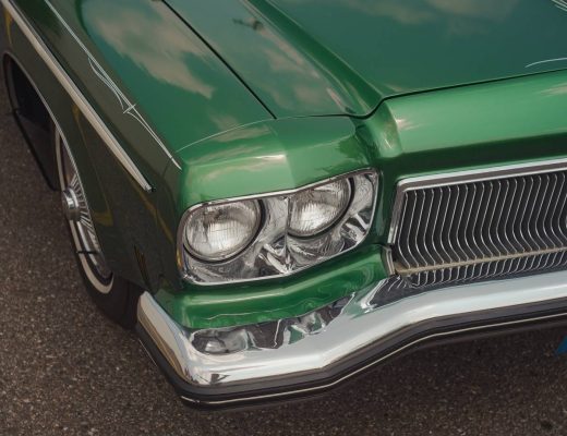 A close-up of a classic green car parked on the pavement. The car's headlights and front-end panel are visible.