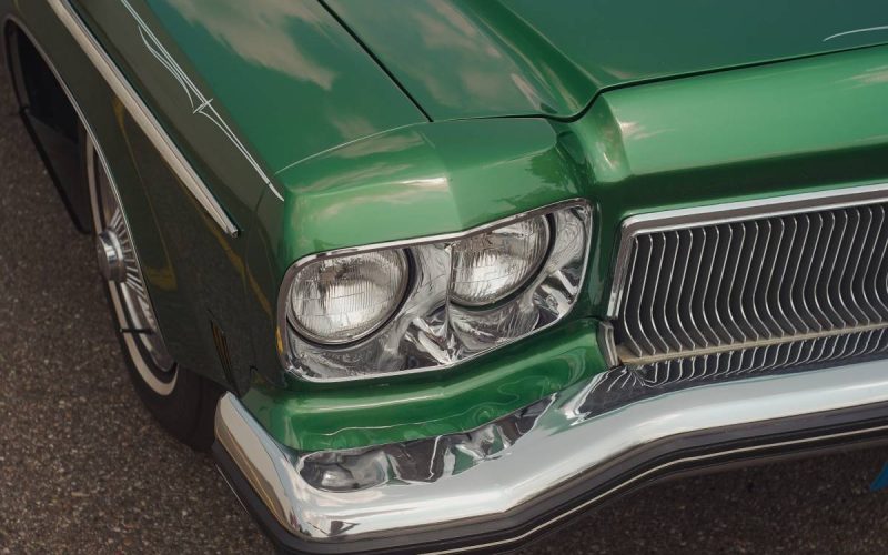 A close-up of a classic green car parked on the pavement. The car's headlights and front-end panel are visible.