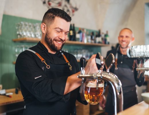 A man pours beer into a glass from a tap. He's smiling and wearing a black apron. Another man is nearby.